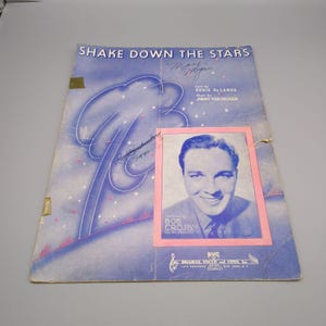 Könnte beinhalten: Vintage Notenblatt mit dem Titel "Shake Down the Stars" mit einem Porträt von Bob Crosby. Das Cover ist blau mit weißen und rosa Akzenten und enthält die Namen des Texters und Komponisten.