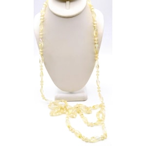 Può includere: Una lunga collana di perline giallo chiaro esposta su un busto bianco. La collana presenta una varietà di forme di perline, tra cui ovali allungati e perline rotonde più piccole. Le perline sono traslucide.