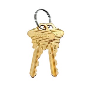 May include: Two gold-coloured keys on a silver key ring. The keys are stamped with "GANNELL SAFE & LOCK" and "USA". The key blades have a notched design. The keys are for a safe or lock.