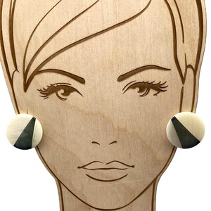 May include: A pair of circular stud earrings featuring a split design with one half in a creamy off-white tone and the other half in a dark charcoal gray. The earrings are displayed on a light-colored wooden cutout shaped like a human face.