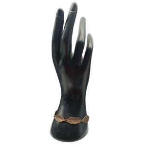 May include: A bronze-colored bracelet featuring a leaf design. The bracelet is displayed on a black hand-shaped jewelry holder. The leaves are connected by small links, creating a delicate and nature-inspired accessory.