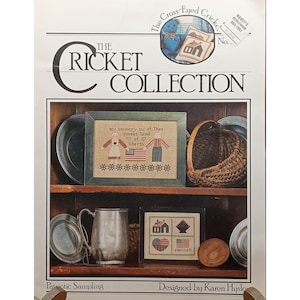 May include: The Cricket Collection patriotic sampling cross-stitch pattern book. The cover features a framed cross-stitch sampler with an American flag and the text "my country tis of Thee sweet land of liberty". Other samplers and a basket are also visible.