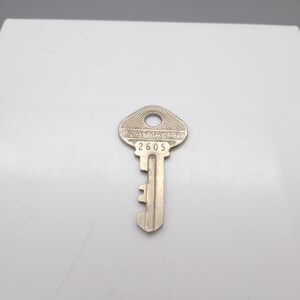 Puede incluir: Una llave plateada con el texto "SLAYMAKER" y el número "2605" estampados en la cabeza. La llave tiene un ojo de cerradura y muescas a lo largo del vástago. La llave está diseñada para una cerradura.