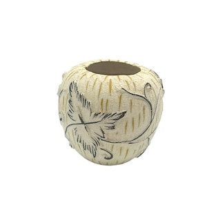 May include: A small, round, off-white ceramic vase with a textured surface. The vase features a leaf and vine design in black and gold. The interior of the vase is brown.