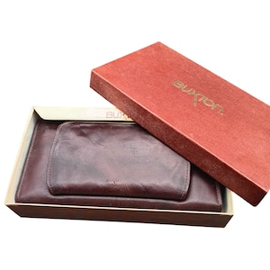 May include: A burgundy leather wallet set, including a larger wallet and a smaller coin purse, presented in a red box with the brand name "Buxton" in gold lettering. The set is displayed open, showcasing the items.
