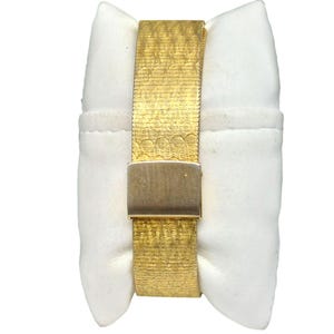 Puede incluir: Un reloj dorado con una banda texturizada y un cierre. El reloj se muestra en un coj&iacute;n blanco.