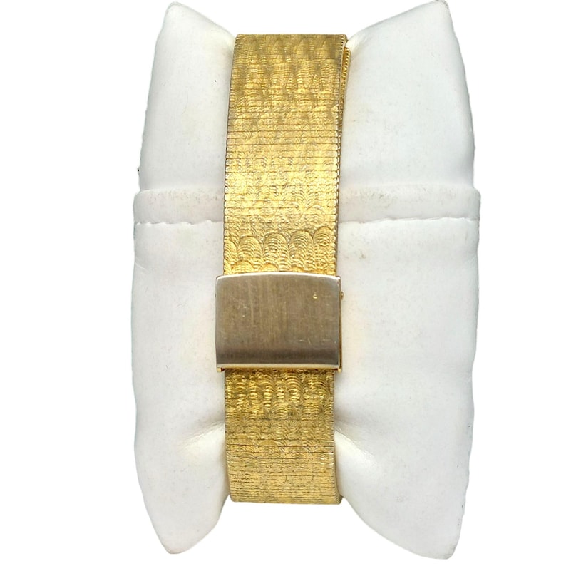 Puede incluir: Un reloj dorado con una banda texturizada y un cierre. El reloj se muestra en un coj&iacute;n blanco.