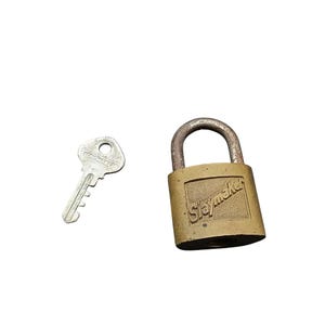 Puede incluir: Un candado de latón antiguo y su juego de llaves. El candado tiene un cuerpo rectangular con la palabra "Slaymaker" grabada en la parte delantera. La llave es de color plateado y tiene la palabra "Slaymaker" en la cabeza.
