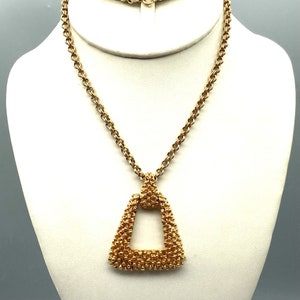 Può includere: Una collana con catena dorata con un grande ciondolo triangolare. Il ciondolo è fatto di piccole perline rotonde e ha un'apertura quadrata al centro.