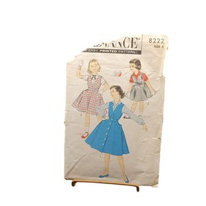 Könnte beinhalten: Vintage Advance Schnittmuster mit Illustrationen von drei Kleidern: ein rot-weiß kariertes Kleid, ein grauer Jumper und ein blaues Knopfkleid. Das Muster ist mit "8222" und "Größe 4" gekennzeichnet. Das Muster ist auf gealtertem Papier gedruckt.