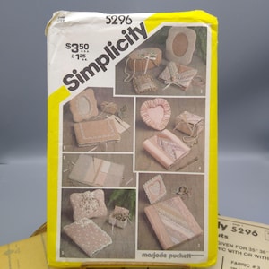 May include: A Simplicity sewing pattern envelope, number 5296, with a yellow and white design. The pattern features various craft projects, including a heart-shaped frame and a small box. The text "marjorie puckett" is visible.