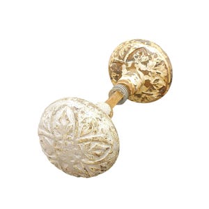 Puede incluir: Un pomo de puerta antiguo con un acabado blanquecino desgastado. El pomo presenta un diseño floral y una varilla metálica con un extremo roscado. La apariencia envejecida sugiere un estilo vintage o rústico, adecuado para la decoración del hogar.