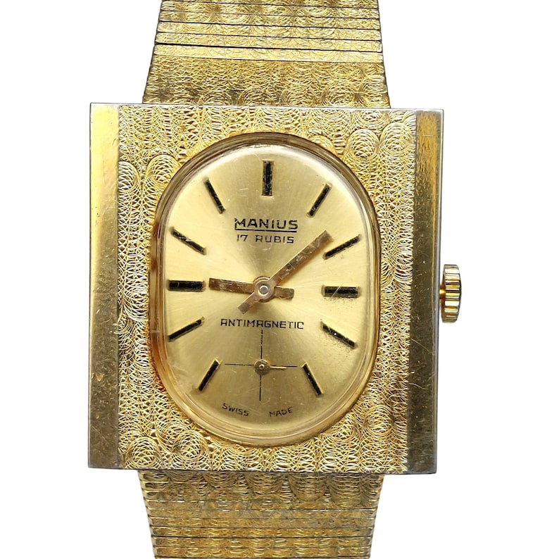 Puede incluir: Un reloj de pulsera dorado con una caja cuadrada y una esfera de forma ovalada. La esfera es dorada con marcas negras y el texto "MANIUS 17 RUBIS" y "ANTIMAGNETIC".