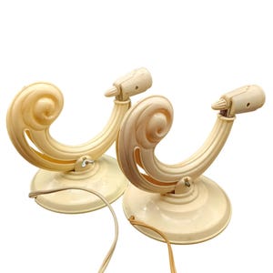 May include: Two vintage off-white wall sconces. Each sconce features a decorative scroll design and a small light fixture. The sconces have a circular base and are connected by electrical cords. The overall design is reminiscent of mid-century decor.