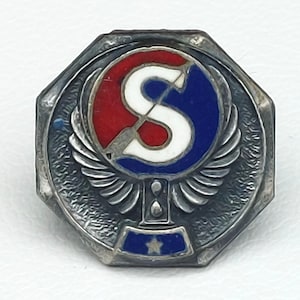 May include: A vintage silver-toned pin with an octagonal frame. The pin features a central design of a white "S" over an arrow, set within a red and blue circle. Below the circle are stylized wings and a blue base with a star.