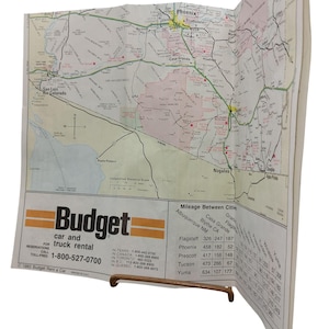 以下が含まれることがあります： 予算レンタカーのロゴが入ったアリゾナ州の折り畳み地図。地図にはフェニックスやツーソンなどの都市が表示され、距離表も記載されています。地図は木製のスタンドで開いた状態に保たれています。