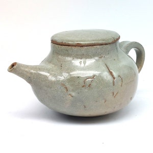 Puede incluir: Una tetera de cerámica gris claro con tapa y un pico. La tetera tiene un aspecto rústico y hecho a mano con un esmalte moteado y marcas marrones.