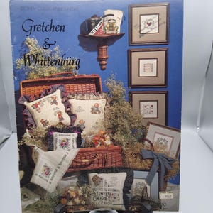 May include: A catalog cover with the text "Gretchen & Whittenburg" and various cross-stitch designs. The image features framed cross-stitch samplers, pillows, and a picnic basket, all showcasing floral and teddy bear motifs. The color palette includes blues, browns, and creams.