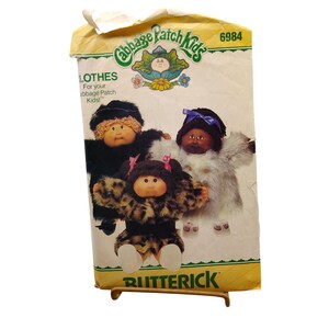 May include: A vintage Butterick pattern for Cabbage Patch Kids clothes. The pattern packet shows illustrations of three Cabbage Patch Kids dolls in various outfits. The packet is yellowed with age and has the number 6984 printed on it.