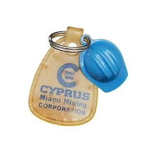 Puede incluir: Un llavero vintage con un dije de casco azul y una etiqueta beige. La etiqueta tiene el texto "CYPRUS Miami Mining CORPORATION" impreso en azul. El llavero está sujeto a un anillo de metal plateado.