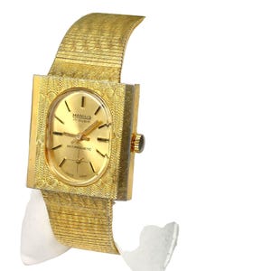 Puede incluir: Un reloj de pulsera dorado con una esfera cuadrada y una correa de metal dorado. La esfera del reloj tiene un fondo blanco con marcadores de hora romanos negros y agujas negras. La esfera del reloj tambi&eacute;n tiene el texto "MANIUS 17 RUBIS" y "ANTIMAGNETIC".