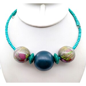 May include: A teal beaded necklace featuring three large, round beads. The outer beads have a marbled design with pink and green accents. The central bead is a solid teal colour, displayed on a white bust.