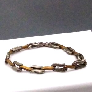 Op de afbeelding: Een zilveren en gouden armband met een kettingschakelontwerp. De schakels zijn gevormd als langwerpige ovalen.