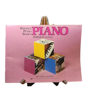 May include: A pink music instruction book titled Bastien Piano Basics Piano Primer Level by James Bastien. The cover features three cubes displaying piano keys and a grand piano, with text printed in black and large pink letters on the front.