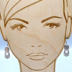 May include: Silver-toned earrings with a spoon-like design, hanging from silver hooks. The earrings are displayed on a wooden stand carved with a face. The earrings have a detailed, vintage aesthetic.