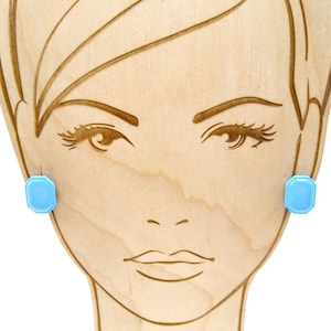 Puede incluir: Expositor de madera con un rostro estilizado con detalles grabados. Pendientes octagonales de color azul claro están sujetos a los lóbulos de las orejas. El rostro tiene pestañas detalladas y una boca sencilla.