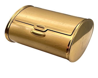 May include: A polished gold-tone metal box with an oval-shaped profile and a hinged lid. The container features a smooth, reflective surface with a central latch mechanism on the front, designed with rounded ends and a curved, cylindrical body shape.