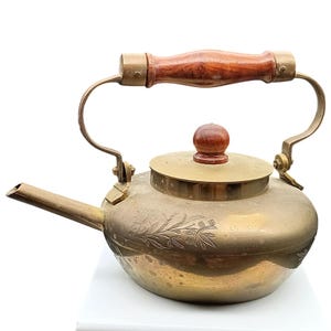 Puede incluir: Una tetera de latón vintage con asa y pomo de tapa de madera. La tetera presenta un pico curvo y grabado floral decorativo. El asa está sujeta con herrajes de latón. La tetera está diseñada para uso en estufa.
