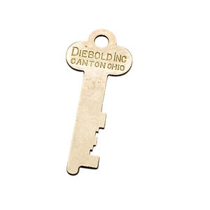May include: A vintage metal key with the text "DIEBOLD INC CANTON OHIO" engraved on the head. The key has a rectangular shaft with several notches cut along one edge. The key is a light, metallic color.