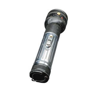Op de afbeelding: Een vintage zaklamp met een zwarte bovenkant en een zilveren, geribbelde behuizing. De zaklamp heeft een schakelaar en een kleine knop aan de bovenkant. Aan de onderkant zit een metalen lus.