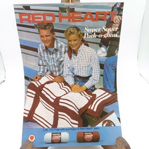 May include: A vintage advertisement for Red Heart Super Saver Pack-a-ghan yarn. The ad features two people sitting on a bleacher with a crocheted blanket in their laps. The text on the ad reads "Red Heart Super Saver Pack-a-ghan" and "L.W. 1117 Free with purchase of Red Heart yarn."