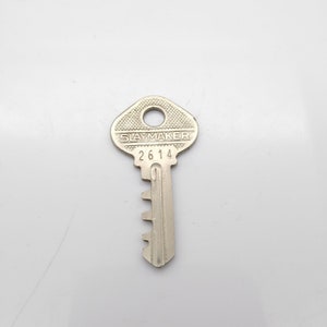 Puede incluir: Una llave plateada con el texto "SLAYMAKER" y el número "2614". La llave tiene un agujero circular en la parte superior y una serie de muescas a lo largo de la hoja. Probablemente es para una cerradura.