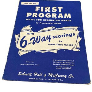 Könnte beinhalten: Ein blaues Musikbuch mit dem Titel First Program Music for Beginning Bands von Prescott und Phillips mit 6-Way scorings von James (Red) McLeod. Das Cover zeigt weißen Text auf blauem Grund mit einem zentralen weißen Grafikelement und Verlagsangaben.