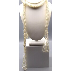 May include: A long, cream-colored pearl necklace displayed on a white jewelry bust. The necklace is draped, with strands of pearls cascading down. The design includes a braided section and beaded tassels, showcasing a classic and elegant style.