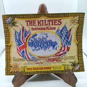 May include: A vintage souvenir album cover featuring a photo of a band in kilts, with the title "The Kilties Souvenir Album" and the text "Songs and Marches of Auld Scotia" printed in bold letters. The cover is decorated with a plaid pattern and images of the American and British flags.