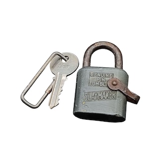 Puede incluir: Un candado Slaymaker gris con una llave. El candado tiene las palabras "GENUINE PIN TUMBLER" y "SLAYMAKER" impresas. La llave tiene un lazo de metal y la palabra "Ilco" impresa en la cabeza.