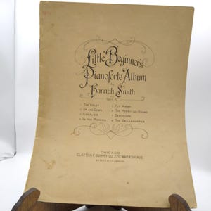 Op de afbeelding: Een vintage bladmuziekcover voor "Little Beginner's Pianoforte Album" van Hannah Smith. De cover is lichtbruin met zwarte tekst en decoratieve krullen. De titel en de naam van de auteur staan prominent weergegeven, samen met een lijst met liedjes.