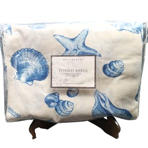 May include: A folded Pottery Barn Tossed Shell tablecloth in a cream color with a blue seashell and starfish print. The product label displays the text "TOSSED SHELL TABLECLOTH" and "70 X 108 INCHES". The item is encased in clear plastic packaging.