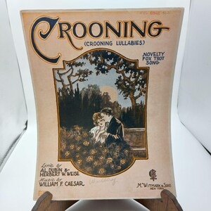 Puede incluir: Una portada de partitura musical vintage para una canción titulada "Crooning" (Crooning Lullabies). La portada presenta a una pareja abrazándose en un entorno de jardín bajo la luna llena. El texto "Novelty Fox Trot Song" está impreso en la esquina superior derecha.