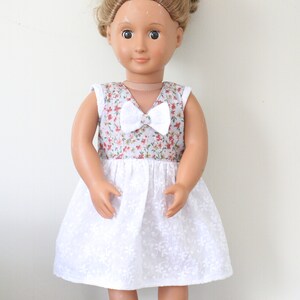 May include: A doll wearing a white dress with a floral print bodice and a white bow at the neckline.
