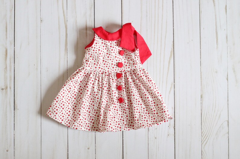 May include: A red and white sleeveless dress with a red bow at the neck and red buttons down the front. The dress is covered in a small red star pattern.