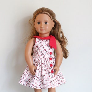 May include: A doll wearing a white dress with red stars and a red bow. The dress has red buttons down the front. The doll has brown hair and brown eyes.