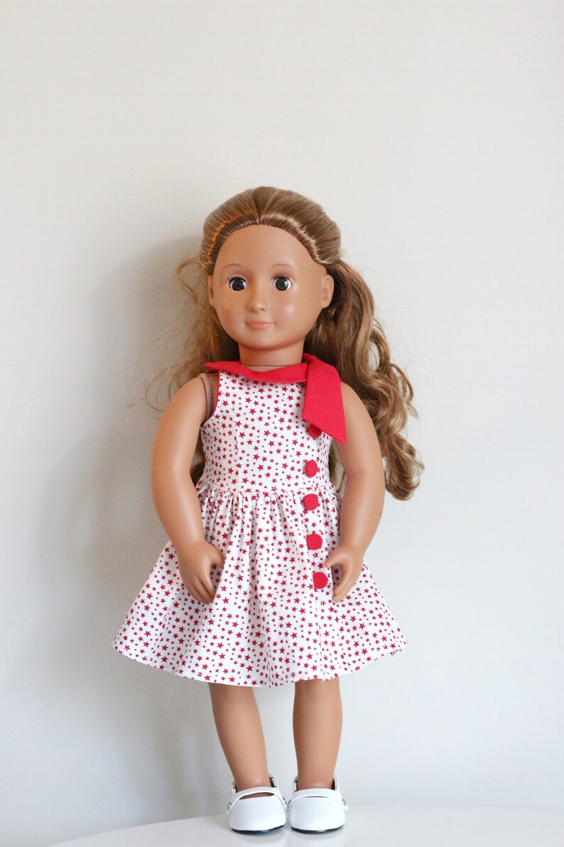 May include: A doll wearing a white dress with red stars and a red bow. The dress has red buttons down the front. The doll has brown hair and brown eyes.
