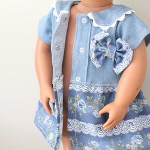 May include: A light blue denim dress with a white lace trim and a floral print. The dress has a bow on the front and a button closure.