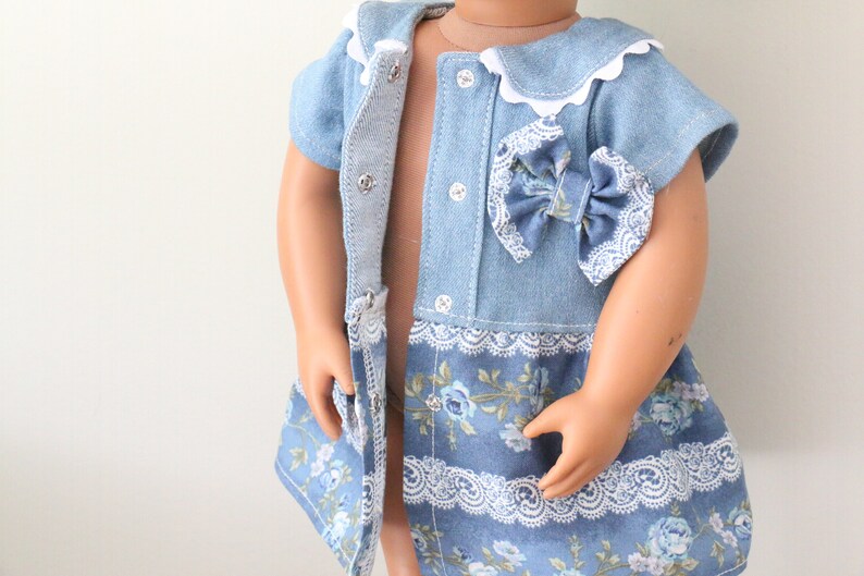 May include: A light blue denim dress with a white lace trim and a floral print. The dress has a bow on the front and a button closure.