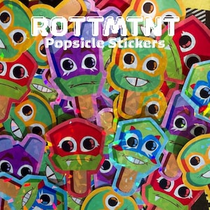 May include: A collection of holographic stickers shaped like popsicles, each featuring a cartoon character face with large eyes and a wooden stick. The text ROTTMTNT Popsicle Stickers is printed in white over the colorful, layered sticker design.
